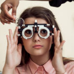 Horizontal shot of interested and curious caucasian girl on appointment with eye care specialist wearing phoropter while ophthalmologist checking her vision, sitting over yellow background.